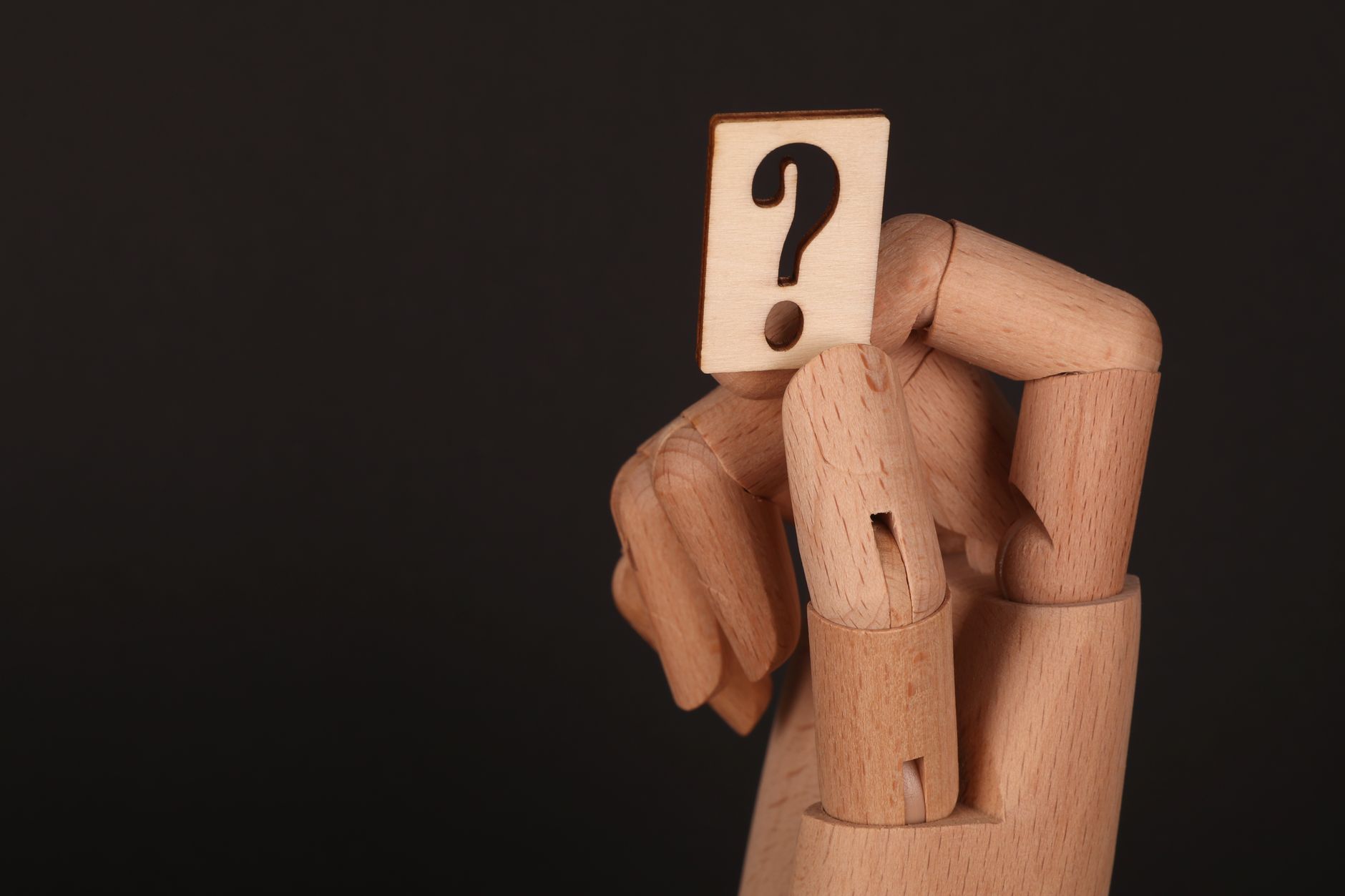 Close-up of a wooden mannequin hand holding a block with a question mark, emphasizing curiosity.
