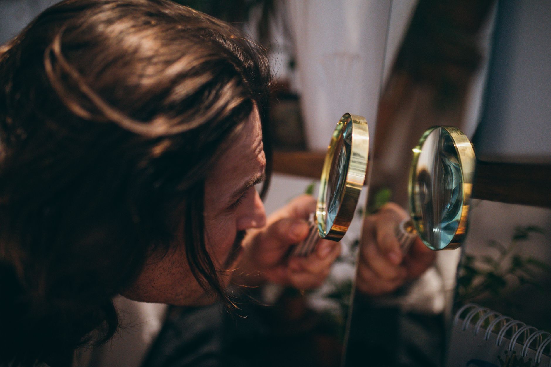 Close-up shot of a man with long hair using a magnifying glass to inspect his reflection in a mirror.
